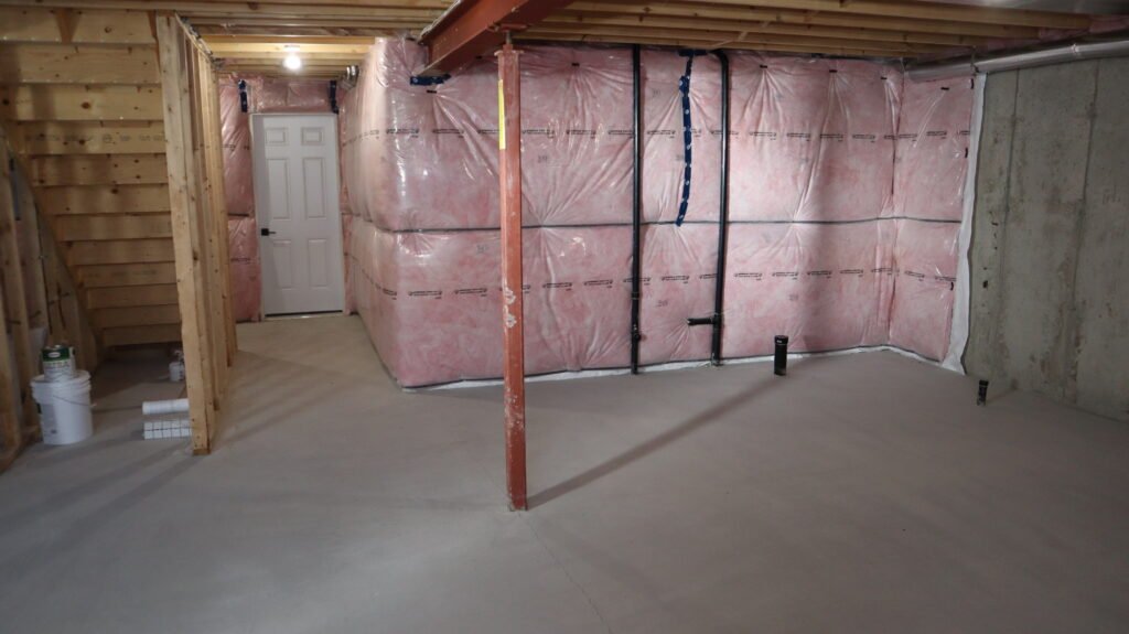 Sam Farmaha at a basement renovation site with pink fiberglass insulation, exposed wooden studs, joists, and visible plumbing pipes along the wall.